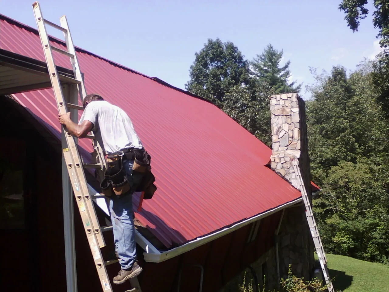 Roofer on a ladder beside a red metal roof and stone chimney — Soffit & Fascia Repair by Roofing Brothers in Oak Run